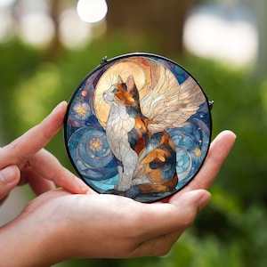 Puede incluir: Una obra de arte circular en vidriera que representa un gato calicó alado, sobre un fondo de luna y cielo estrellado. El gato es en tonos naranja, blanco y marrón, con un fondo azul y dorado. La pieza se sostiene en las manos de una persona.