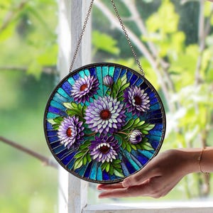 May include: Round stained glass window art featuring a bouquet of purple and white flowers with green leaves. The flowers have red centers and are set against a blue and turquoise background. The artwork is framed in black and hangs from a chain.