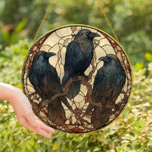 May include: A round stained glass window featuring three black crows perched on a brown branch. The crows have detailed feathers and are set against a cream and brown background with a gold border. The window is hanging from a gold chain.