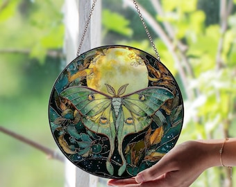 May include: Round stained glass window art featuring a large green moth with yellow accents, set against a full moon and foliage. The artwork is framed in black and hangs from a chain, creating a decorative piece.