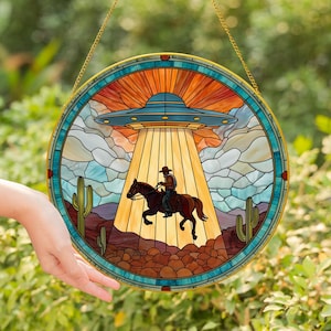 May include: Round stained glass window art featuring a cowboy on horseback being abducted by a UFO. The design includes a blue and orange sky, a desert landscape with cacti, and a gold-colored border. The window is hanging from a gold chain.
