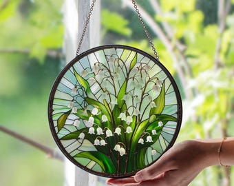 May include: Round stained glass window panel featuring a lily of the valley design. The artwork showcases white bell-shaped flowers with green leaves against a light green and blue background. The panel is framed with a dark metal border and hangs from a chain.