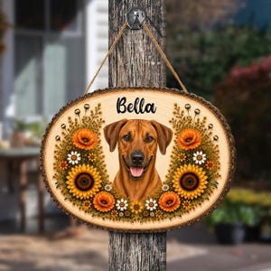 May include: An oval wooden plaque with bark edges, hanging by a jute rope. It features a central illustration of a brown dog with a black muzzle and the name Bella printed above. The dog is framed by orange sunflowers and small white flowers.