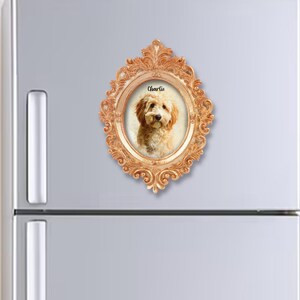 May include: An ornate, gold-coloured oval frame with a portrait of a dog named Charlie. The dog has light brown fur and is centered within the frame. The frame is attached to a refrigerator. The frame has decorative detailing.