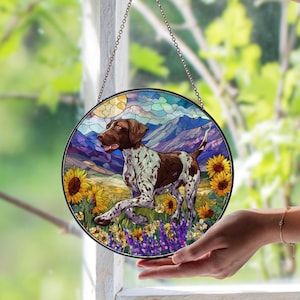 May include: Round stained glass window art featuring a brown and white dog in a field of sunflowers and lavender. The background includes mountains, a sun, and a blue sky. The artwork is framed with a black metal border and a hanging chain.