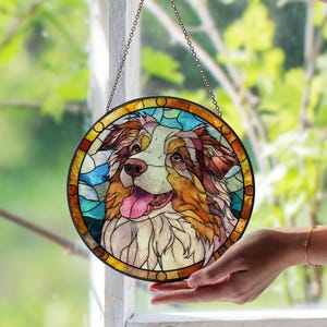 May include: Circular stained glass window depicting an Australian Shepherd dog. The artwork uses shades of brown, white, and tan. The dog's tongue is pink. The window is framed in black and has a hanging chain.