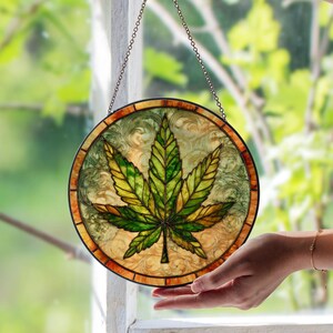 Op de afbeelding: Rond gebrandschilderd glazen raam met een cannabisbladontwerp. Het blad is uitgevoerd in groen- en geeltinten, met gedetailleerde nerven. Het raam heeft een decoratieve rand en wordt opgehangen aan een ketting.