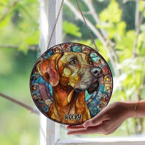 Könnte beinhalten: Ein runder Glasfenster-Anhänger mit einem Hundepotrait. Der Hund ist in Braun-, Orange- und Gelbtönen dargestellt, mit einem blauen und mehrfarbigen Hintergrund. Der Name "Rocky" steht unten. Er wird von einer Metallkette gehalten.
