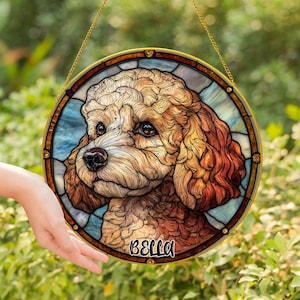 May include: Round stained glass window featuring a detailed portrait of a dog with brown and tan fur. The dog's name, "BELLA," is written in black script at the bottom. The window has a gold-colored frame and is suspended by a gold chain.