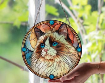 May include: Round stained glass window art featuring a cat with blue eyes and brown and white fur. The cat is centered in a circular design with a stained glass effect, with blue and orange accents. The window is hanging from a chain.