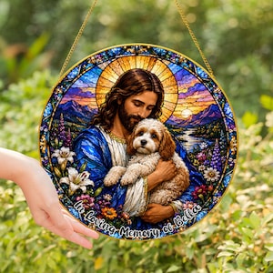 May include: A circular stained glass suncatcher featuring a bearded figure in blue robes holding a small brown and white puppy. The background depicts a sunset landscape with mountains and flowers. The text In Loving Memory of Bella is printed.