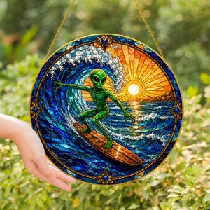 May include: A circular stained glass suncatcher featuring a green alien surfing on a wooden surfboard. The scene depicts a large blue ocean wave, white sea foam, and a bright yellow sun setting over the horizon, all framed by a decorative border.