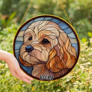 May include: A round stained glass window featuring a detailed portrait of a dog with brown and tan fur. The dog's name, "Rocky," is written in white script at the bottom. The window is framed in gold and hangs from a gold chain.