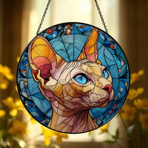 Puede incluir: Vidriera redonda que representa un gato Sphynx con ojos azules. La cabeza del gato está representada en tonos beige, naranja y rosa, sobre un fondo azul con elementos decorativos. Una cadena negra está unida para colgar.