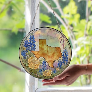 Pode incluir: Arte de janela redonda em vitral com o contorno do Texas em tons de ouro e castanho. O design inclui flores azuis, rosas amarelas e outros elementos florais. A janela é suspensa por uma corrente.