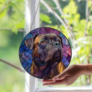 Peut inclure: Pièce d'art en vitrail rond représentant un portrait de chien. L'œuvre utilise un style mosaïque avec des nuances de marron, de violet et de bleu. Une chaîne en métal est attachée pour l'accrocher. La pièce est tenue devant une fenêtre.