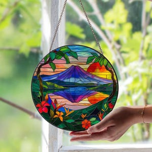 May include: Round stained glass panel depicting a mountain landscape with a lake and sunset. The artwork features vibrant colors, including purple, blue, orange, and green. The panel is framed with a black border and hangs from a chain.