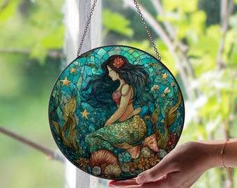 Puede incluir: Vidriera redonda que representa una sirena con largo cabello negro y una flor roja. La sirena está sentada entre algas marinas, conchas y estrellas de mar. La obra de arte está en tonos turquesa, verde y naranja, con un marco de metal negro y una cadena para colgar.
