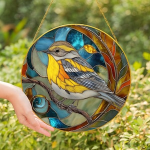 May include: A round stained glass window featuring a colorful bird perched on a branch. The bird has yellow, gray, and blue feathers. The background is a mix of blue, green, and orange glass, with a gold-colored metal frame and hanging chain.