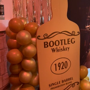 May include: A brown cardboard cutout of a whiskey bottle with the text "BOOTLEG Whiskey 1920 SINGLE BARREL PROHIBITION BLEND". The cutout is next to a wall decorated with gold balloons.