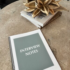 Könnte beinhalten: Ein graues spiralgebundenes Notizbuch mit dem Text "INTERVIEW NOTES" auf dem Einband. Das Notizbuch liegt auf einem Tisch mit einem Stapel Bücher und einer Trockenblumen-Arrangement.