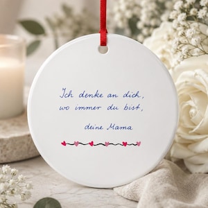 Puede incluir: Un adorno redondo de cerámica blanca que cuelga de una cinta roja. El adorno presenta un texto azul en cursiva que dice: Ich denke an dich, wo immer du bist, deine Mama. Debajo del texto hay una línea horizontal con pequeños corazones rosas y rojos.