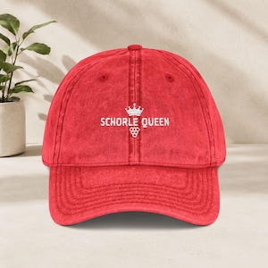 Op de afbeelding: Een rode honkbalpet met een verweerde, verwassen textuur. De voorkant is voorzien van wit geborduurde tekst "SCHORLE QUEEN", geplaatst onder een klein kroonicoontje en boven een afbeelding van een tros druiven.