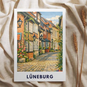 Könnte beinhalten: Ein rechteckiger Kunstdruck mit einer farbenfrohen Illustration einer gepflasterten Straße in Lüneburg, Deutschland. Die Szene zeigt Fachwerkhäuser aus Backstein, schwarze Wandlaternen und bunte Blumen. Das Wort LÜNEBURG ist unten aufgedruckt.