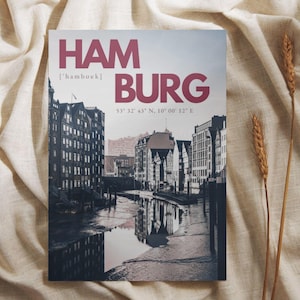 Könnte beinhalten: Ein rechteckiger Wandkunstdruck mit einem Schwarz-Weiß-Foto von Hamburger Gebäuden entlang eines Kanals. Der Text HAMBURG ist oben in großen dunkelrosa Buchstaben gedruckt, darunter die phonetische Schreibweise und die Koordinaten 53° 32' 43" N, 10° 00' 12" E.