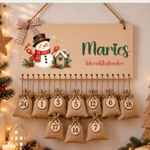 May include: A rectangular wooden advent calendar sign featuring a snowman illustration, a plaid bow, and the text Maries Adventskalender. Below the sign, multiple small burlap drawstring bags hang from twine, each with a numbered wooden tag.