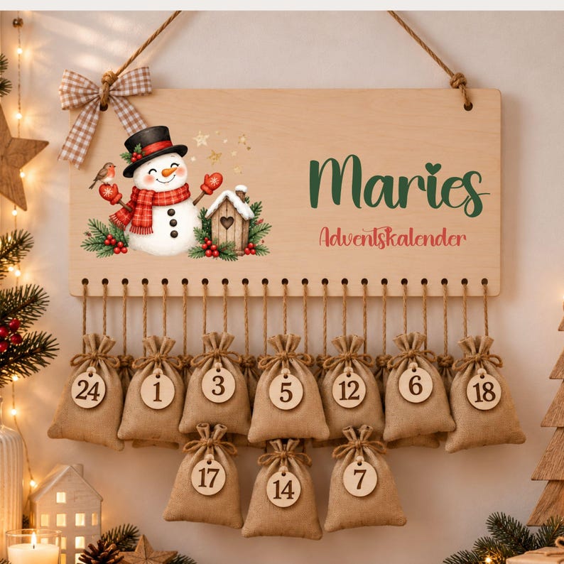 May include: A rectangular wooden advent calendar sign featuring a snowman illustration, a plaid bow, and the text Maries Adventskalender. Below the sign, multiple small burlap drawstring bags hang from twine, each with a numbered wooden tag.