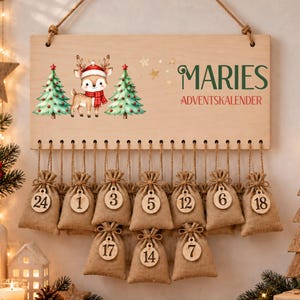 May include: A rectangular wooden advent calendar sign featuring a cartoon deer in a red hat and scarf between two green Christmas trees. The text reads MARIES ADVENTSKALENDER. Below, ten small burlap drawstring bags hang from twine with wooden number tags.