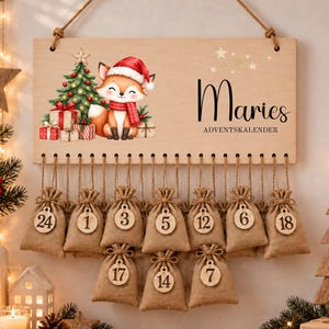 May include: A rectangular wooden wall hanging advent calendar featuring a cartoon fox in a Santa hat, a Christmas tree, and gifts. The text reads Maries ADVENTSKALENDER. Ten small burlap drawstring bags with numbered wooden tags hang below.