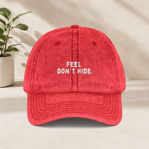 Könnte beinhalten: Eine rote Baseballkappe mit verwaschener, abgenutzter Textur. Auf der Vorderseite der Kappe befindet sich der gestickte weiße Text "FEEL DON'T HIDE.". Die Mütze hat eine strukturierte Krone, einen gebogenen Schirm und einen Knopf oben.