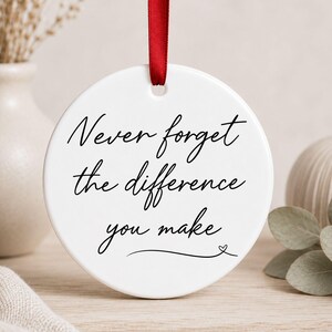 Könnte beinhalten: Ein rundes, weißes Keramikornament mit dem schwarz aufgedruckten Text Never forget the difference you make und einem kleinen Herzsymbol am Ende. Das Ornament ist an einem dünnen roten Band befestigt, das durch ein Loch oben verläuft.