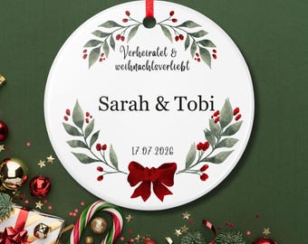 May include: A round white ceramic ornament featuring a red ribbon hanger. The design includes a watercolor wreath of green leaves and red berries, with the text Verheiratet & weihnachtsverliebt, the names Sarah & Tobi, and the date 17.07.2026.