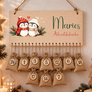 May include: A rectangular wooden advent calendar sign with a hanging rope, featuring two illustrated penguins in winter hats and scarves, the name Maries, and the text Adventskalender. Ten small burlap drawstring bags with numbered wooden tags hang below.