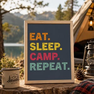 Op de afbeelding: Een ingelijst bord met de woorden "EAT. SLEEP. CAMP. REPEAT." in oranje, geel, rood en turquoise. Het bord staat op een houten oppervlak met een kampeermok en lantaarn op de achtergrond. De mok heeft de tekst "THE ADVENTURE BEGINS."
