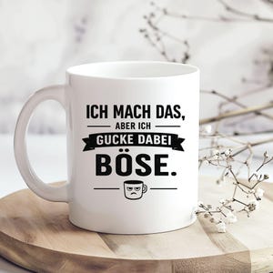 Könnte beinhalten: Eine weiße Keramiktasse mit dem schwarzen Text "ICH MACH DAS, ABER ICH GUCKE DABEI BÖSE." und einer kleinen Grafik eines grimmig schauenden Tassengesichts. Das Design enthält horizontale Linien und ein Banner-Element auf der Vorderseite.