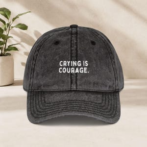 Könnte beinhalten: Eine anthrazitfarbene Baseballkappe mit verwaschener, abgenutzter Textur. Auf der Vorderseite befindet sich der weiß gestickte Text "CRYING IS COURAGE.". Die Kappe hat einen gebogenen Schirm, einen Knopf oben und genähte Ösen zur Belüftung.
