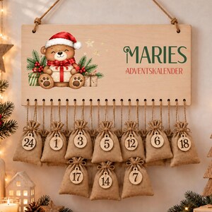 May include: A rectangular wooden advent calendar sign featuring a cartoon teddy bear in a Santa hat holding a gift, with the text MARIES ADVENTSKALENDER. Twenty-four small burlap drawstring bags with numbered wooden tags hang from the sign.