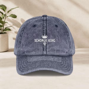 Pode incluir: Um boné de beisebol azul-marinho com efeito lavado, aba curva e tira ajustável. A parte frontal apresenta o texto bordado em branco SCHORLE KING, posicionado abaixo de um ícone de coroa e acima de um ícone de cacho de uvas.