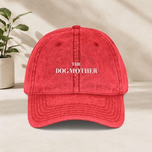 Könnte beinhalten: Eine rote Baseballkappe mit einer verwaschenen, abgenutzten Textur. Auf der Vorderseite der Kappe ist der weiße Schriftzug "THE DOGMOTHER" in einer klaren serifenlosen Schrift eingestickt. Die Kappe hat eine strukturierte Krone und einen Schirm.