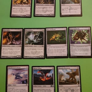 May include: A collection of Magic: The Gathering cards featuring various creatures and artifacts. The cards are displayed on a green surface. The cards include Tangle Hulk, Bladed Sentinel, Sultai Banner, Dross Ripper, Gust-Skimmer, Maze Skullbomb, Dross Skullbomb, Thundersteel Colossus, Meteor Golem, and Dromoka Monument.