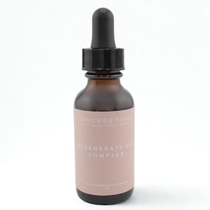 May include: A brown glass bottle with a black dropper. The bottle has a pink label with the text "SPACE23 SKIN" and "REGENERATE OIL COMPLEX". The bottle contains a clear liquid.