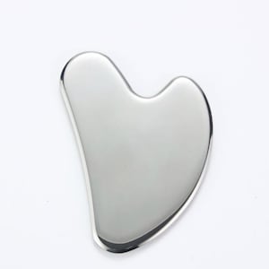 Op de afbeelding: Een gepolijst, zilverkleurig gua sha-gereedschap. Het gereedschap heeft een gladde, gebogen vorm met een hartvormige inkeping aan de bovenkant. De gua sha is ontworpen voor gezichtsmassage en huidverzorgingsroutines. Het gereedschap is gemaakt van een reflecterend metaal.