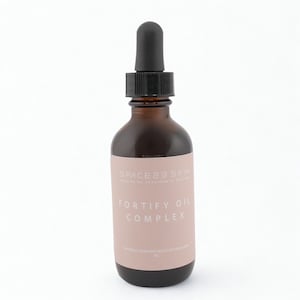 May include: A brown glass bottle with a black dropper. The label displays "SPACE23 SKIN" and "FORTIFY OIL COMPLEX" in white text on a pink label. The bottle contains oil.