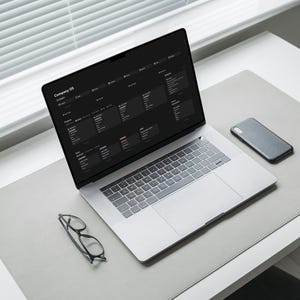 May include: A laptop displaying a dark-themed interface, a smartphone, and eyeglasses rest on a gray desk mat. The laptop screen shows the text "Company OS". The scene is illuminated by natural light from a nearby window.
