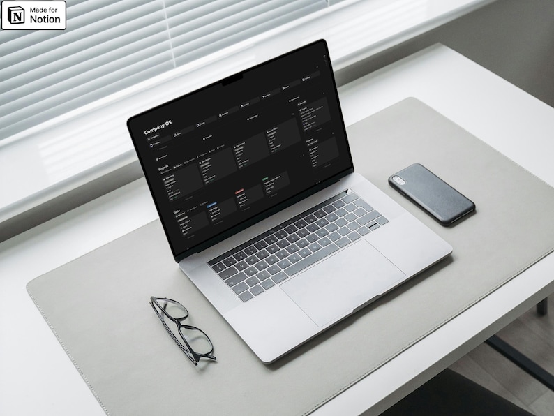 May include: A laptop displaying a dark-themed interface, a smartphone, and eyeglasses rest on a gray desk mat. The laptop screen shows the text "Company OS". The scene is illuminated by natural light from a nearby window.