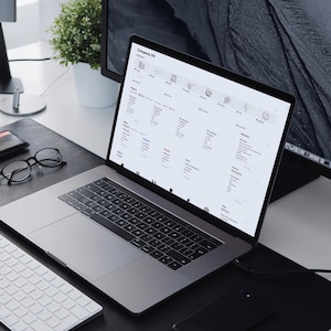 May include: A silver laptop displaying a webpage with a grid of icons and text sits on a black desk. A white keyboard, glasses, wallet, and phone are also on the desk. A monitor and plant are in the background. The text "Made for Notion" is in the top left corner.